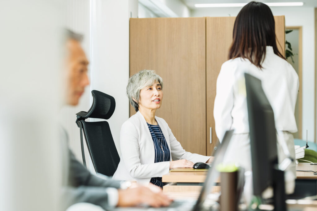 オフィスで女性管理職が部下と向き合って話をしている様子。職場での業務調整や相談の場面