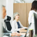 オフィスで女性管理職が部下と向き合って話をしている様子。職場での業務調整や相談の場面